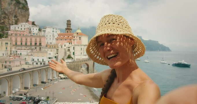 Smiling woman pointing at Amalfi Coast town taking selfie video for social media