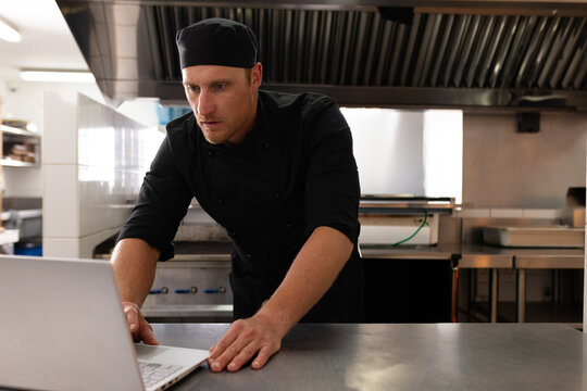 Male chef leaning over stainless steel countertop using laptop in kitchen with gas stove and hood
