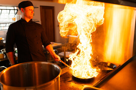 Male chef flipping flaming pan in restaurant kitchen, with stock pot and exhaust hood, copy space