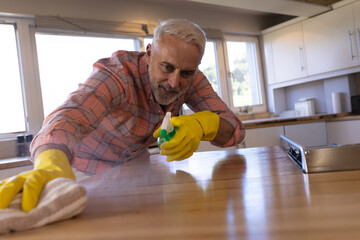 Senior man spraying cleaning solution and wiping wooden island in modern kitchen with yellow gloves
