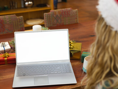 Woman sitting in living room wearing Santa hat, looking at laptop with wrapped gifts