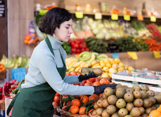 Adult woman seller in apron puts fresh pears on display in vegetable shop