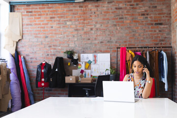 Young adult woman talking on smartphone using laptop in fashion studio with fabrics, copy space