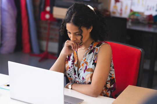 Young woman leaning forward at white desk in creative studio, using silver laptop and notebook
