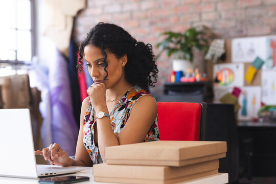 Woman working on laptop in creative studio while sorting mailer boxes smartphone sketches swatches