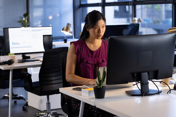 Asian woman working on computer at open-plan office, with laptop keyboard mouse and succulent plant