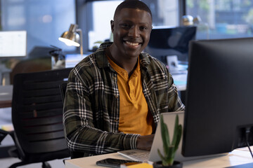 African American man smiling while using laptop at open-plan office, with monitor and succulent