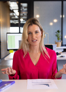 Woman speaking, gesturing at open-plan office desk, with printed bar chart sheet and purple book