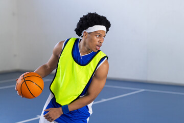 Teenage African American boy dribbling orange basketball in gym wearing green pinnie, copy space