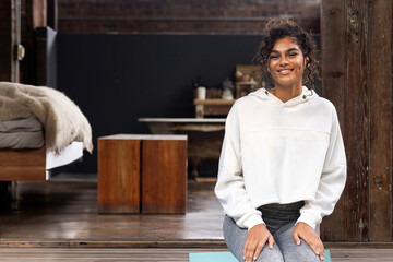 Multiracial woman sitting on teal yoga mat in loft style room, smiling at camera, copy space