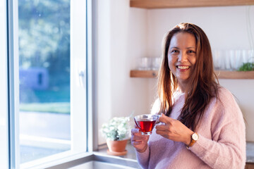 Asian woman standing in kitchen by window holding glass cup of red tea with potted plant