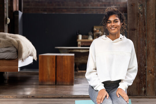Multiracial woman sitting on teal yoga mat in loft style room, smiling at camera, copy space