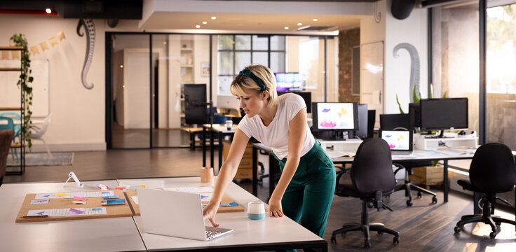 Woman standing at central table in office, working on laptop with color swatches, sticky notes