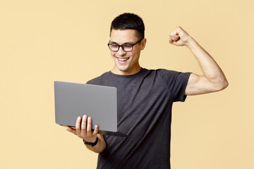 Online win, success at work and great offer. Happy young blogger guy in glasses holds laptop and expresses emotions of victory, isolated on light sand background, empty space, studio shot