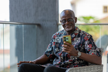 Senior African citizen uses his smartphone for social media and video calls his family outside the...