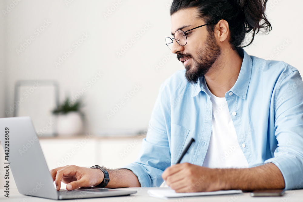 Poster young indian man entrepreneur using laptop and taking notes while working in modern office, sitting 