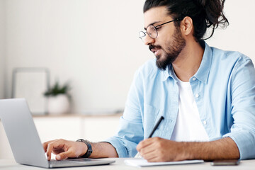Young indian man entrepreneur using laptop and taking notes while working in modern office, sitting at desk and looking at computer screen, wester freelancer guy writing in notepad, copy space