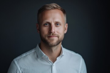 Fototapeta premium Portrait of a young Caucasian man with blue eyes and a beard, wearing a white shirt against a dark background.