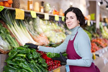 Adult woman seller in apron puts fresh bell peppers on display in vegetable shop
