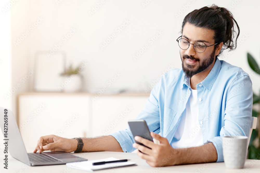 Poster millennial arab freelancer guy using smartphone while working on laptop at desk in home office, brow