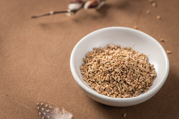 Fennel seeds in a white bowl on a textured brown surface with a delicate feather and blurred plant stem in the background