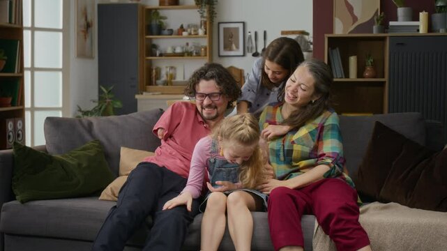 Medium shot of cheerful Caucasian husband with limb difference disability, loving wife, teenage son and daughter sitting on couch, cuddling, playing tickling game, pet maltipoo dog jumping around