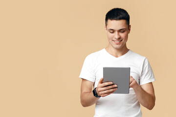 Modern work and leisure time with mobile gadget. Attractive friendly european young guy blogger using digital tablet, surfing in internet, isolated on beige background, studio shot, free space