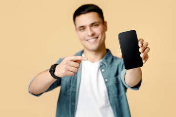It is cool, best mobile app and digital ad. Happy young man in casual shows finger at smartphone with blank screen, selective focus on device, isolated on beige background, studio shot, free space