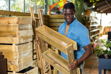 African american gardener, standing in a warehouse in a flower greenhouse, attentively examines a rack of pallets for planting flower sprouts