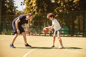 Obraz premium Sports Concept. Side view portrait in action of man and boy playing basketball together on the field