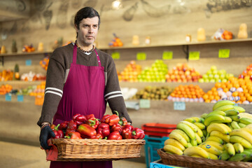 Adult man seller in apron with basket of fresh bell peppers in vegetable shop