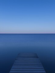 Fototapeta premium Serene wooden dock extending into a calm, deep blue lake under a clear blue sky. Peaceful, minimalist landscape photography.