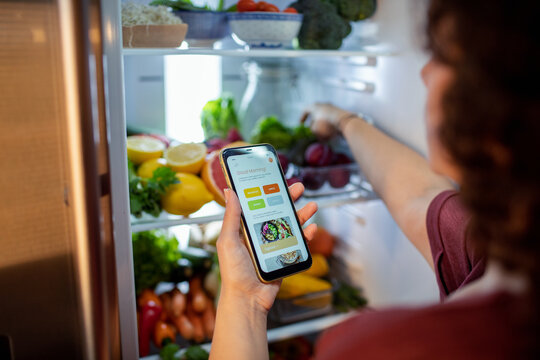 Middle aged mixed ethnicity woman using a dieting app on her smart phone while looking through her fridge
