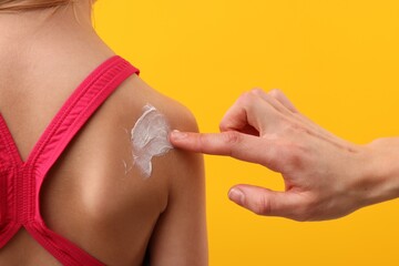 Mother applying sun protection cream onto her daughter's skin on yellow background, closeup