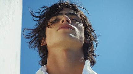 Sun-kissed face tilted upwards, eyes closed against a vibrant blue sky. Windblown dark hair and warm skin tones create a serene, summery mood