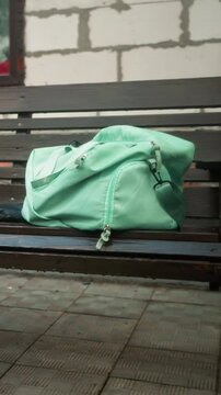 Mint-colored bag and blue umbrella resting on wooden swing bench outside, with background showing partially constructed building, greenery, and interlocked pavement