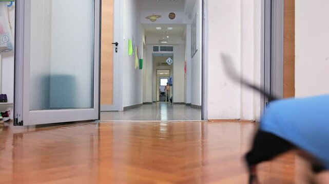 A small black dachshund wearing a blue and white costume walks toward the camera down a brightly lit hallway with polished floors and open office or clinic, school doors