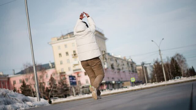 Female figure swinging arms and stepping gracefully across snowy urban plaza, wearing white puffer jacket, beanie and headphones, expressing emotion through fluid dance under pale sky