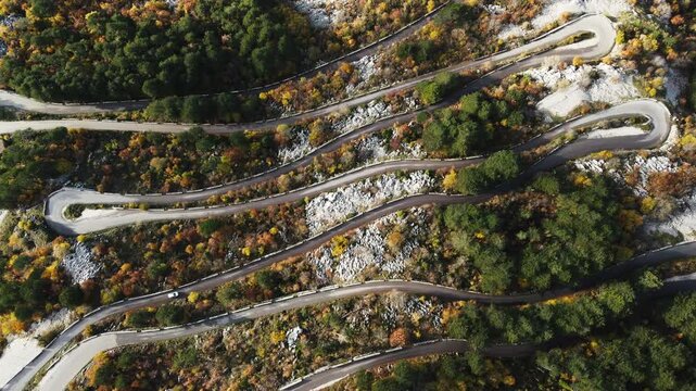 A car winds along a serpentine road through a breathtaking mountain landscape, captured from above. The aerial view reveals mountains draped in rich green moss, highlighting the serene power and