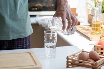 Close up hands of active strong man pouring fresh pure water from pitcher into glass in morning, male drink mineral natural in cup after wake up for health and wellbeing in kitchen in house.