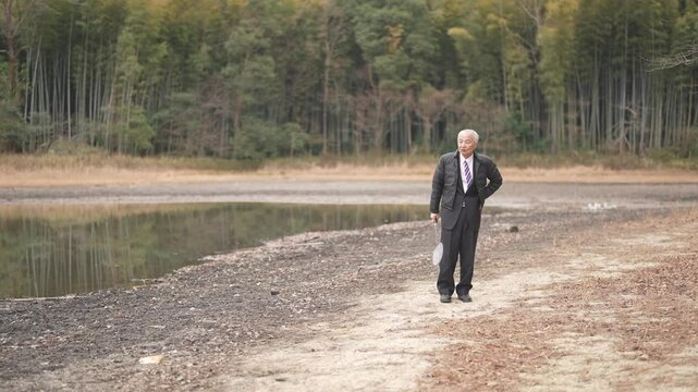 The bamboo forest pond at noon in spring in Japan has plenty of greenery. Japanese men in the late 70s stand in suits and down jackets. He plays badminton with his son in his 30s.