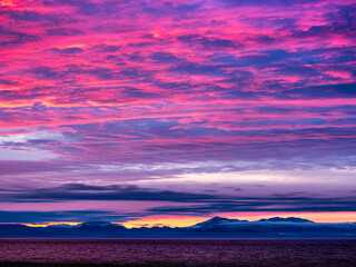 海岸沿いから半島を望む、空には厚い雲に覆われているが夕焼けの陽差しで赤く染まる、手前は海で遠くには半島の山並みが連なっている横構図です。