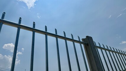 Security Fence Under Blue Sky: A close-up view of a sturdy metal fence against a vibrant blue sky dotted with fluffy white clouds. The image evokes feelings of security, protection and peace of mind.