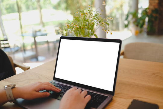 Person using smartphone and laptop with blank screens on wooden table near window in cafe, ideal for app or UI mockup.