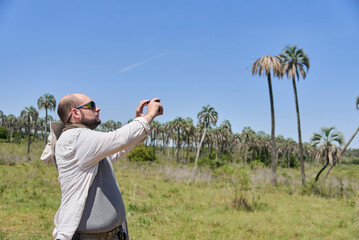 Man traveler taking a picture with his cell phone of the palm tree landscape in front of him to...