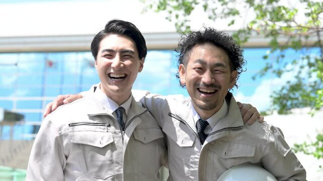 青空の中作業着姿でビルの前でカメラ目線の施工管理などの作業者のイメージ　カメラ目線で肩を組むの動画　スロー