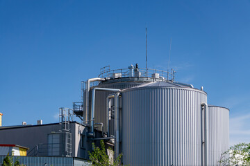 an industrial complex dominated by a large, cylindrical metal tank is visible. The structure is surrounded by technical infrastructure, such as pipes, railings and stairs. The facility is located unde