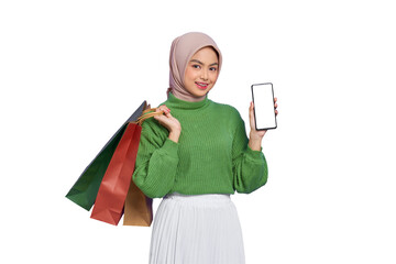 Cheerful beautiful Asian woman in green sweater holding shopping bags, showing blank screen mobile phone isolated on transparent background