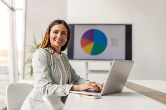 Portrait of skilled female manager sitting at corporate office and typing data on laptop while smiling at the camera.