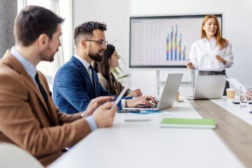 A businessman sitting at boardroom surrounded by coworkers and taking notes on laptop during the presentation.